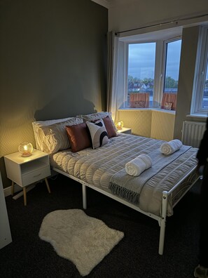 A neatly made bed with a patterned bedspread and pillows, a bedside lamp, and a view of the outdoors through a window.