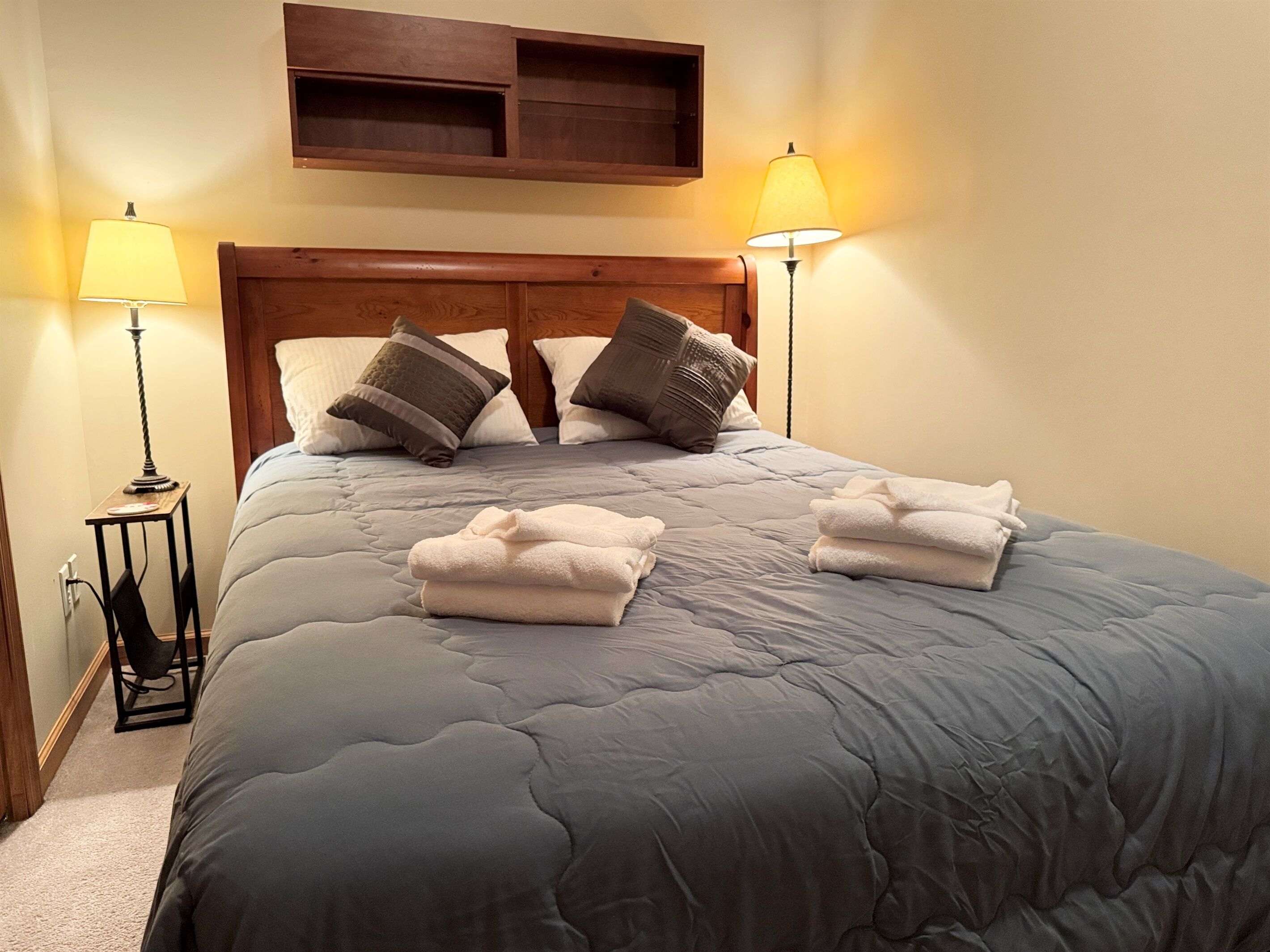 Una cama bien tendida con edredón gris, dos lámparas de noche y un cabecero de madera.