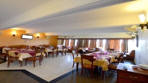A hotel dining area with multiple tables and chairs, a television mounted on the wall, and a large window with curtains. - Barbet Tourist Resort (Sohna)