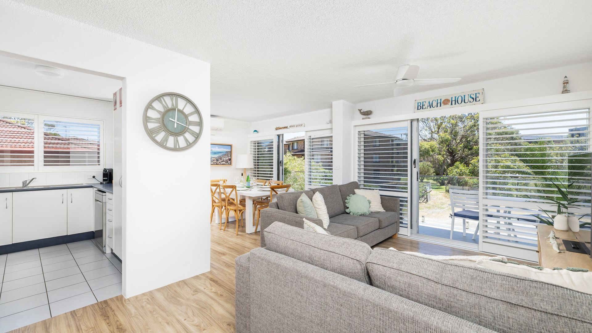 A modern living room with a grey sofa, a dining area with a table and chairs, a kitchen with white cabinets, and a large window with blinds.