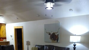 Un salon avec un canapé en cuir noir, un fauteuil blanc, une table basse et un tapis. Un tableau représentant un taureau est accroché au mur, ainsi qu’une lampe et un ventilateur de plafond.
