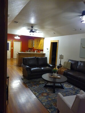 A living room with a black leather sofa, a coffee table, and a patterned rug. There is a wooden floor and a kitchen in the background.