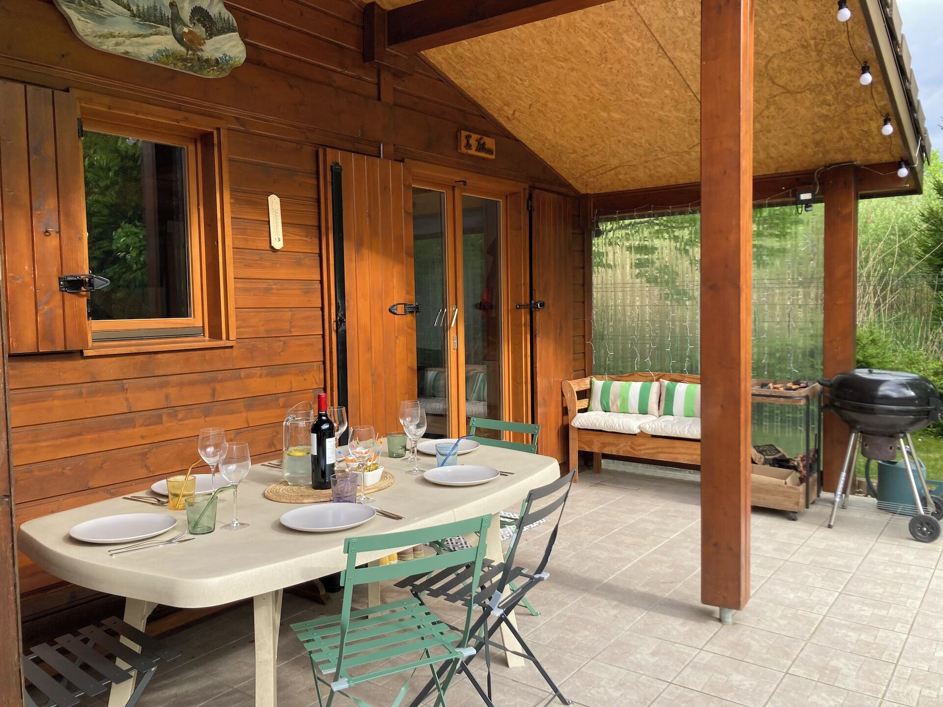A wooden cabin with a covered patio, a dining table set with plates, glasses, and a bottle of wine, and a grill in the background.