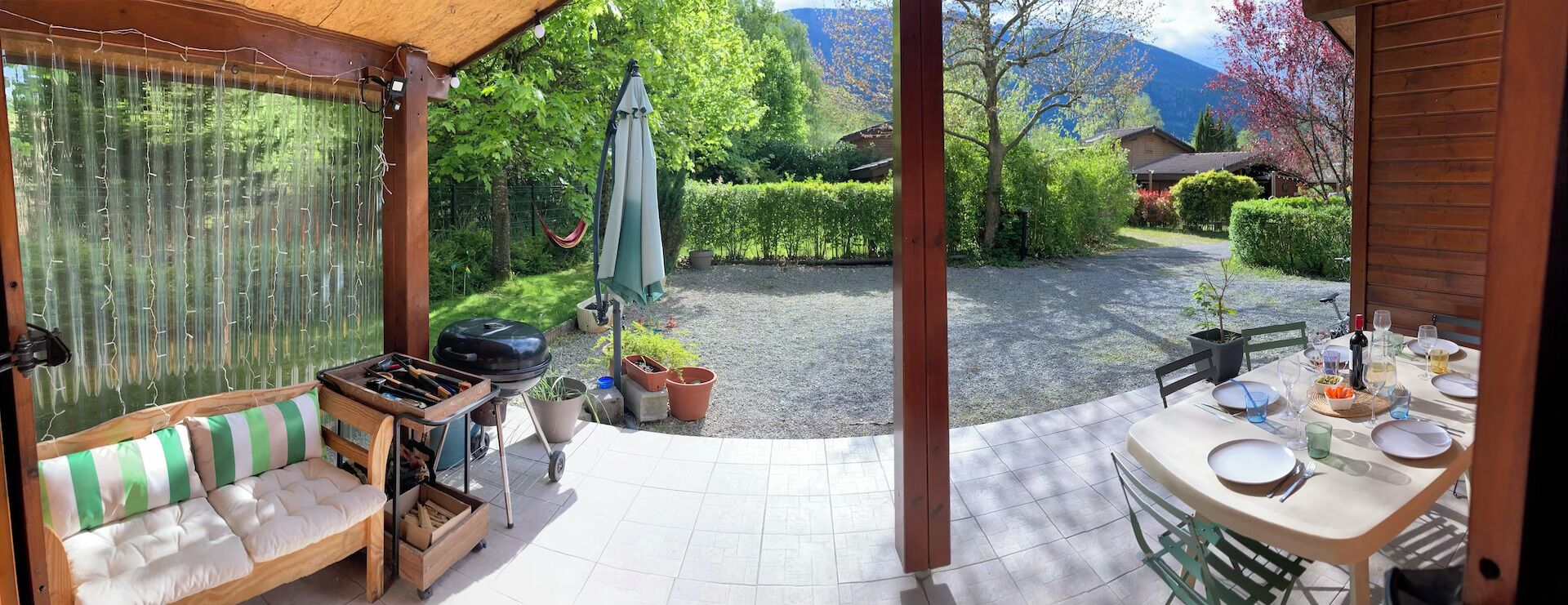 Une terrasse couverte équipée d’un barbecue, de sièges et d’une table dressée pour les repas, donnant sur un jardin avec vue sur une montagne.