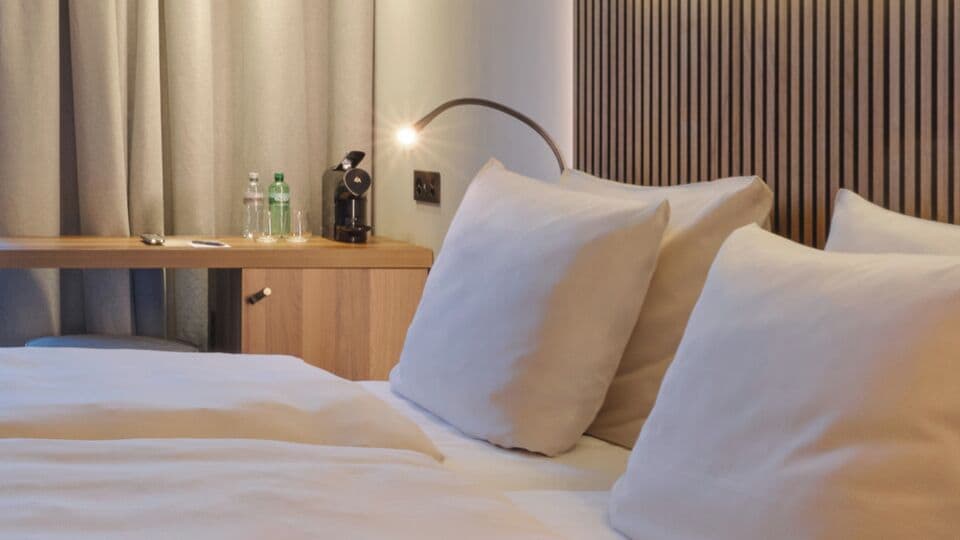 A hotel room with a bed, pillows, a desk with a lamp, and a minibar.