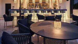 An outdoor bar area with round tables and chairs, a well-lit bar counter with shelves of bottles, and hanging pendant lights. - Dorint Resort & Spa Locarno Riazzino (Locarno)