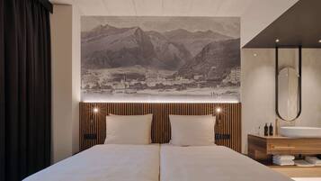 Quarto de hotel com uma cama, uma grande tapeçaria na parede representando uma paisagem montanhosa, cabeceira de madeira, mesa de cabeceira com espelho e banheiro moderno com pia e porta-toalhas.