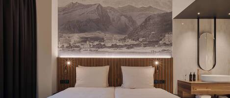 Une chambre d’hôtel comprenant un lit, une grande tapisserie murale représentant un paysage montagneux, une tête de lit en bois, une table de chevet avec un miroir et une salle de bain moderne équipée d’un lavabo et d’un porte-serviettes.