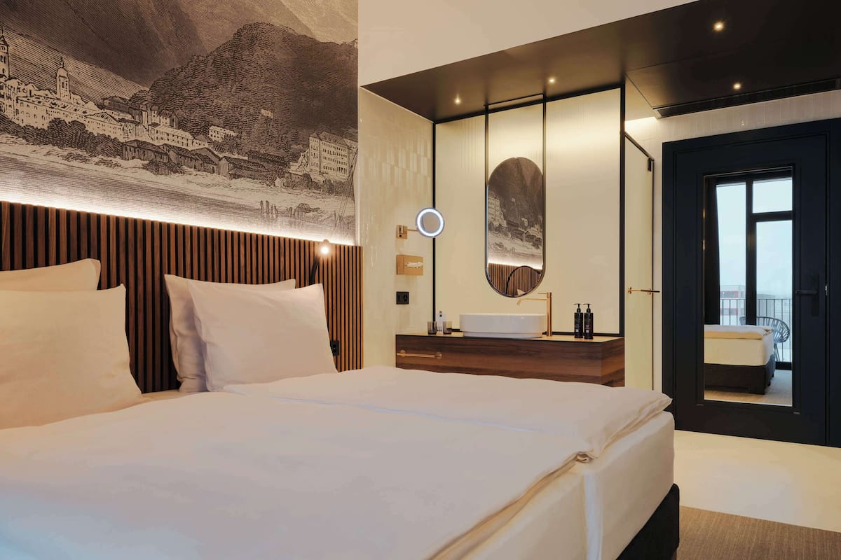 A modern hotel room with a large bed, a wooden headboard, a round mirror, a vanity with a sink, and a view of the city through a large window.