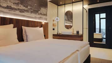 Ein modernes Hotelzimmer mit einem großen Bett, einem hölzernen Kopfteil, einem runden Spiegel, einem Waschtisch und Blick auf die Stadt durch ein großes Fenster.