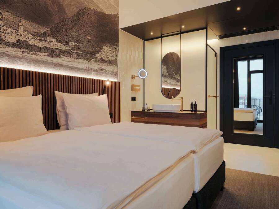Ein modernes Hotelzimmer mit einem großen Bett, einem hölzernen Kopfteil, einem runden Spiegel, einem Waschtisch und Blick auf die Stadt durch ein großes Fenster.