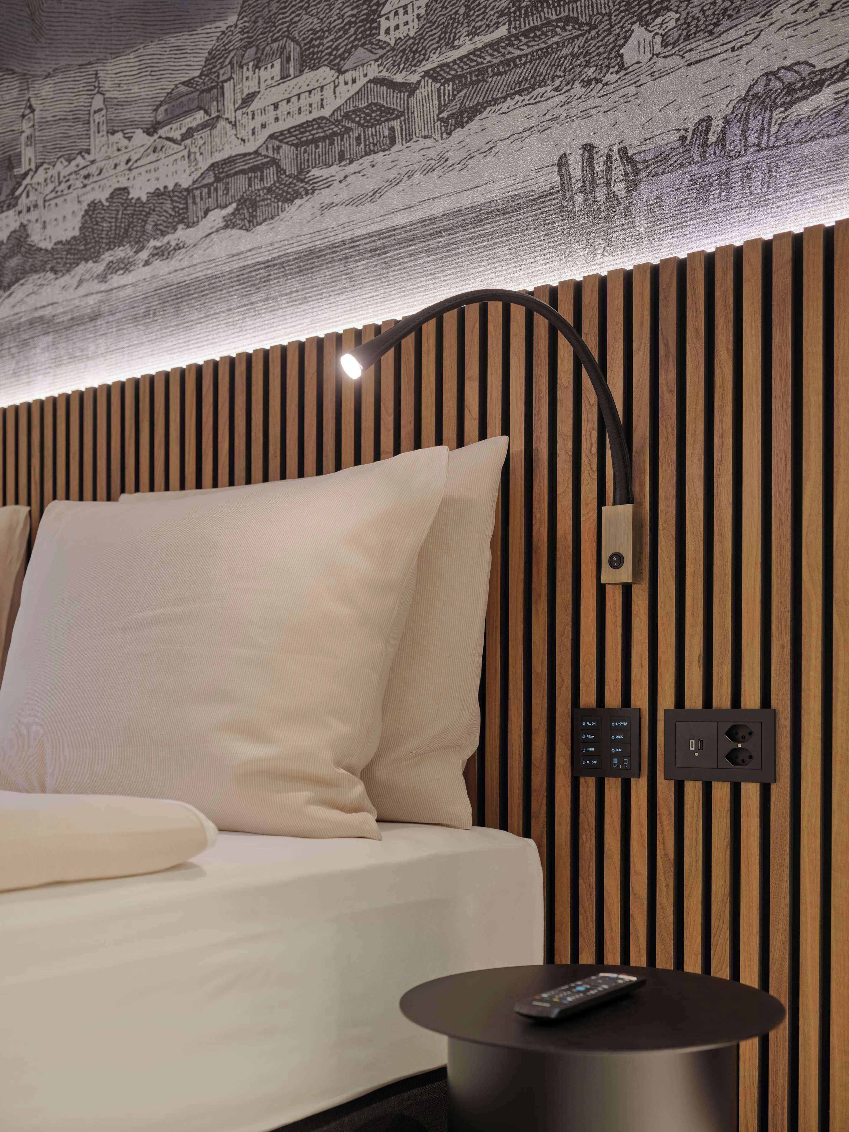 A hotel room with a bed, a bedside table with a remote control, a wall-mounted light, and a power outlet.