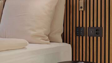 A hotel room with a bed, a bedside table with a remote control, a wall-mounted light, and a power outlet.
