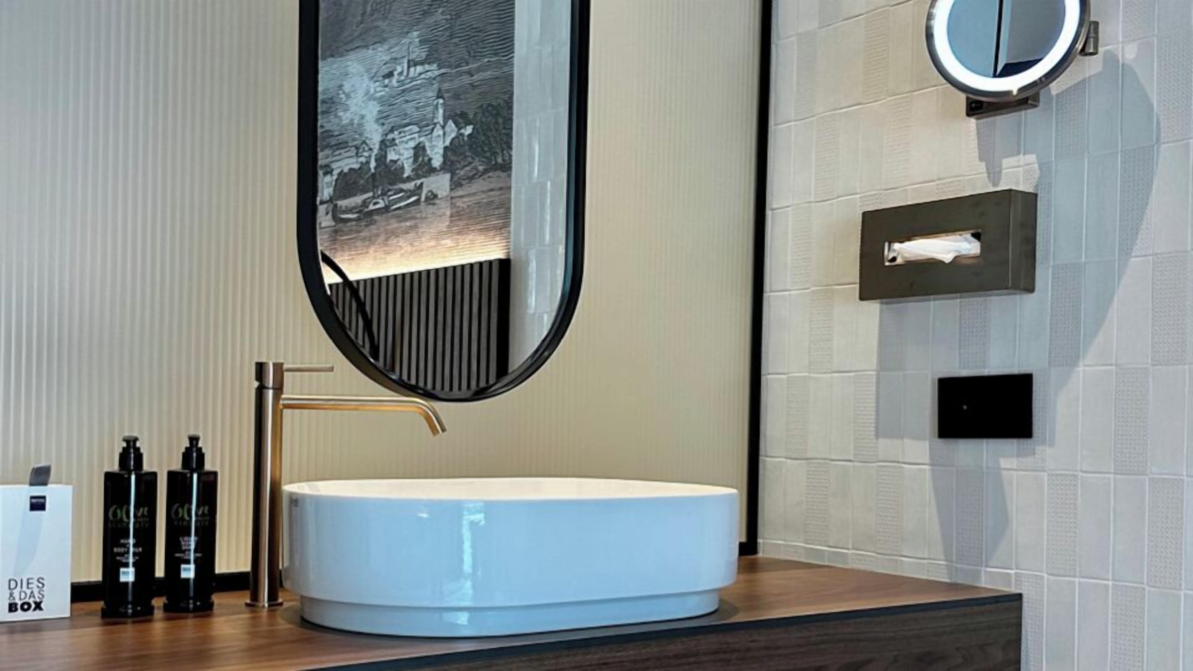 Un baño moderno con un lavamanos blanco redondo, un espejo grande y un dispensador de toallas.