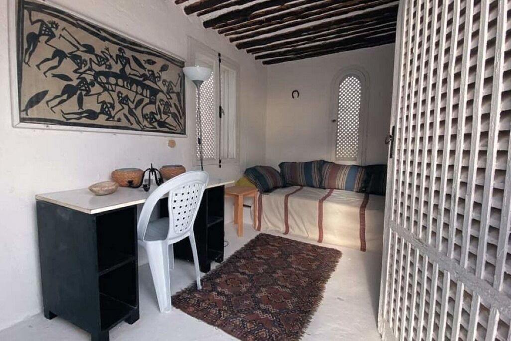 A small, cozy room with a desk, chair, and a sofa. There is a large painting on the wall, a patterned rug on the floor, and a lattice screen on one side.