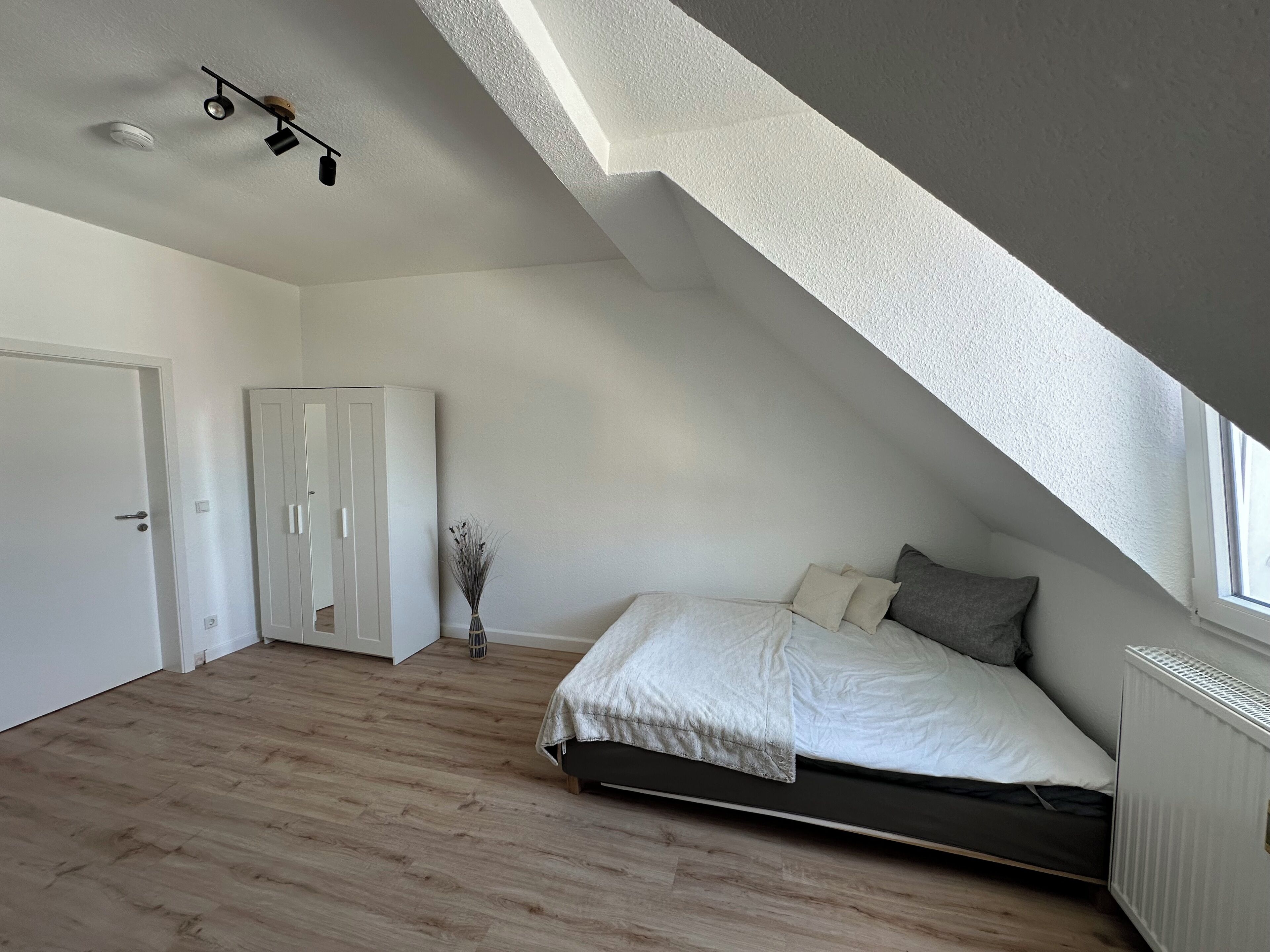 A bedroom with a sloped ceiling, a bed with white and gray bedding, a radiator, and a wooden floor.