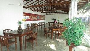 A spacious indoor area with wooden tables and chairs, a large painting on the wall, and a potted plant. - Trinco Rest House (Trincomalee)