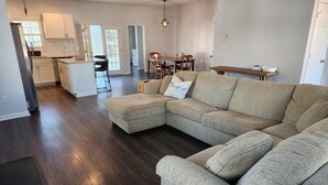 A spacious living room with a sectional sofa, a dining area with a wooden table, and a kitchen with white cabinets.