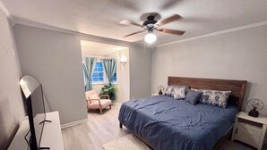 Een slaapkamer met een bed, een plafondventilator, een nachtkastje, een lamp en een stoel.
