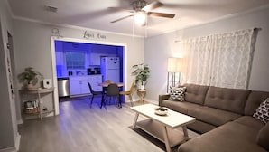 Een woonkamer met een bruine bank, een salontafel en een plafondventilator. Achterin is een keuken zichtbaar met blauwe verlichting.