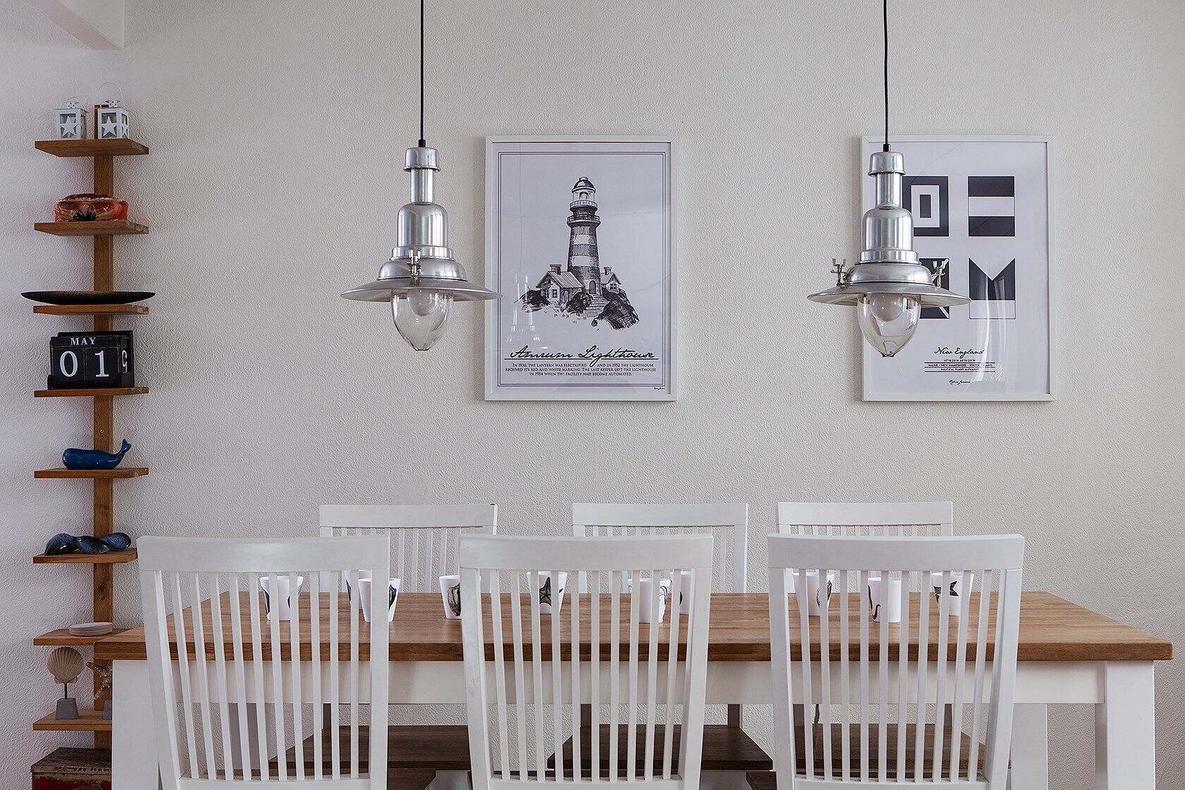 A dining area with a wooden table and white chairs, two pendant lights, a wall-mounted clock, and two framed pictures.