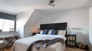 A bedroom with a large bed, a dark headboard, a nightstand with a lamp, and a window with a view of trees. - Semesterboende på Öland (Kalmar län)