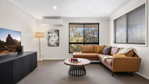 A modern living room with a sectional sofa, a round coffee table, a flat-screen TV on a cabinet, and a large window with blinds. - NorthStar 2, perfectly situated in the heart of Jindabyne (Jindabyne)