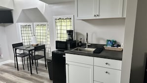 A compact kitchen with a black refrigerator, a microwave, a coffee maker, and a sink. There is a dining table with chairs and a window with plaid curtains.