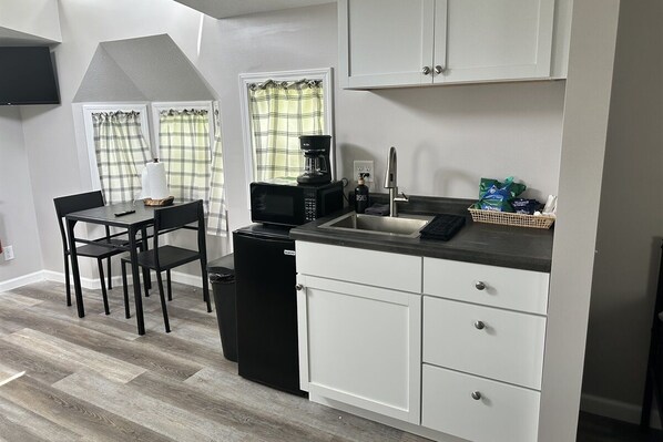 A compact kitchen with a black refrigerator, a microwave, a coffee maker, and a sink. There is a dining table with chairs and a window with plaid curtains.