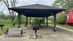 A gazebo with benches and picnic tables, surrounded by a grassy area and trees.