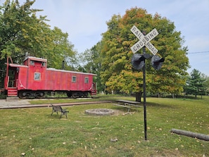 A red caboose train car on tracks with a railroad crossing sign and a bench in a grassy area. - Train caboose with open floor plan, high ceilings & modern airy feel (Hanna City)