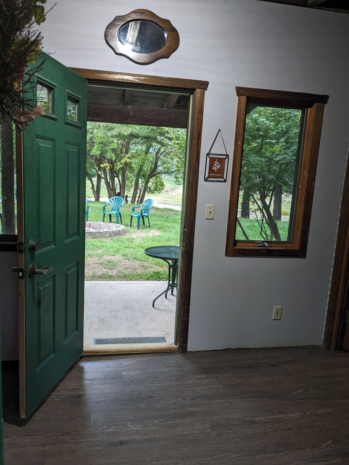 An open door leading to an outdoor area with a table and chairs, a window with a view of trees, and a wall-mounted picture frame.