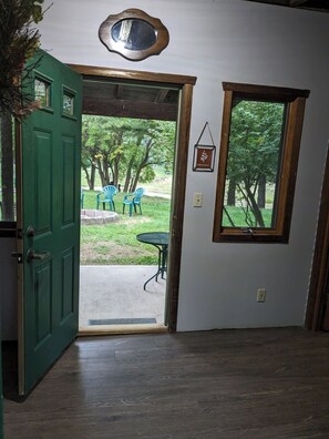 An open door leading to an outdoor area with a table and chairs, a window with a view of trees, and a wall-mounted picture frame.