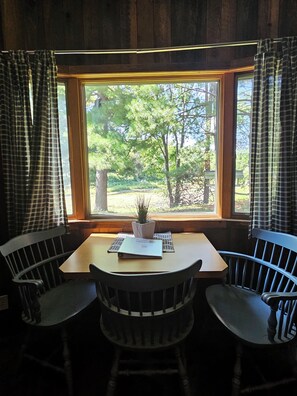 A room with a wooden table and two chairs, a window with a view of trees, and a potted plant on the windowsill.