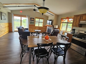 Un coin repas avec une table et des chaises en bois, un ventilateur de plafond, et une cuisine équipée d’une cuisinière et de placards.