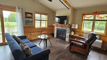 A cozy living room with a fireplace, wooden walls, a ceiling fan, and a large window.