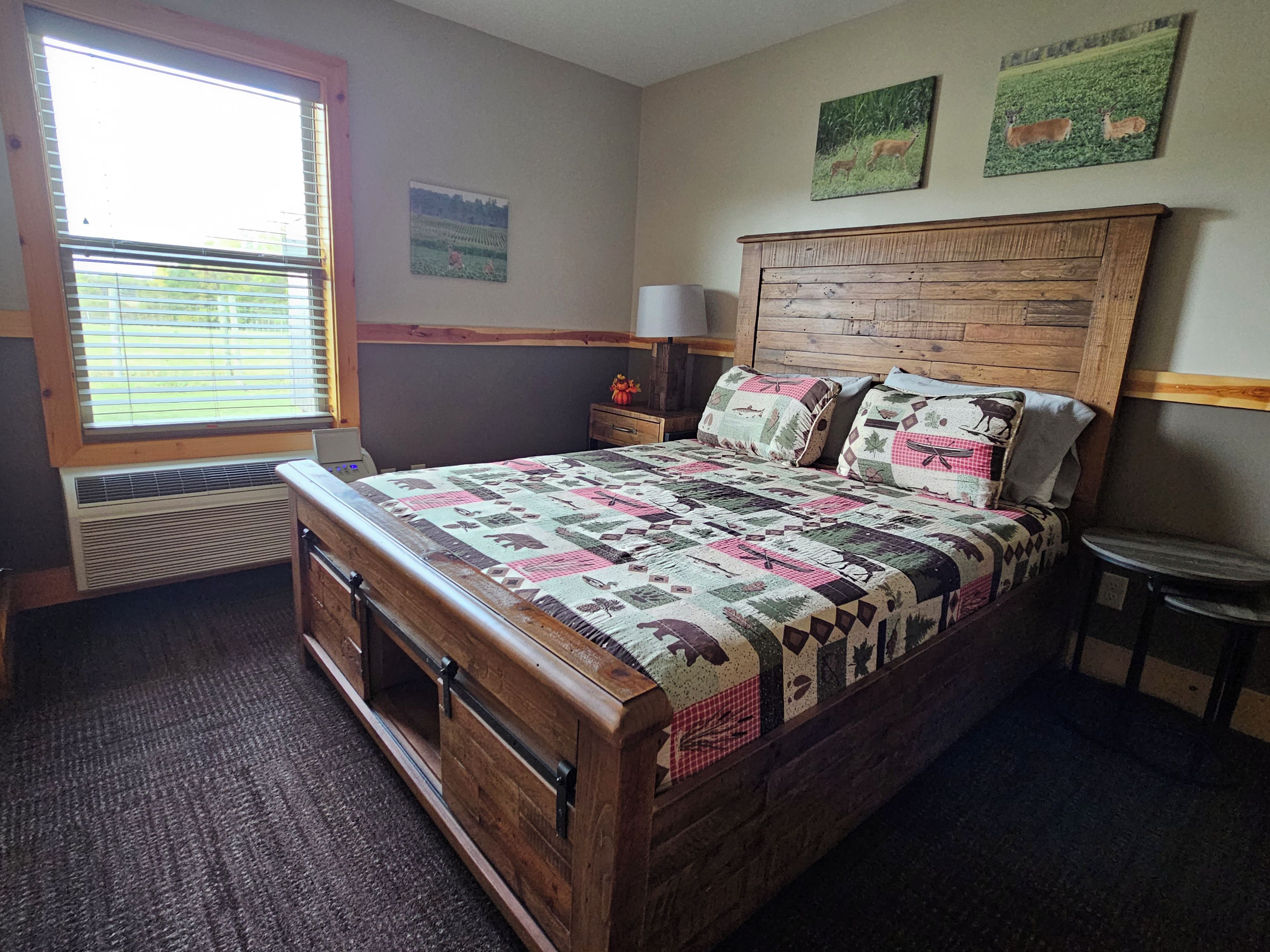 A bedroom with a wooden bed, a window with blinds, a nightstand, a lamp, and two wall-mounted pictures.