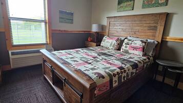 A bedroom with a wooden bed, a window with blinds, a nightstand, a lamp, and two wall-mounted pictures.