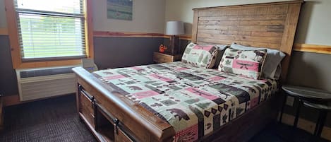 A bedroom with a wooden bed, a window with blinds, a nightstand, a lamp, and two wall-mounted pictures.