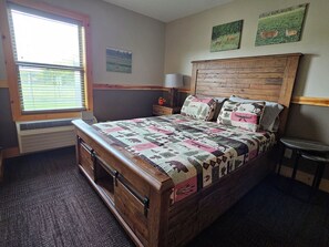 Une chambre à coucher avec un lit en bois, une fenêtre avec des stores, une table de chevet, une lampe et deux tableaux accrochés au mur.