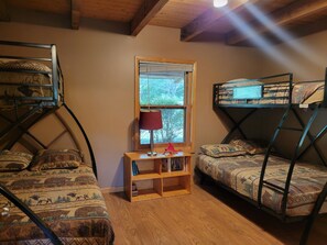 A dormitory room with bunk beds, a window, a wooden shelf with books, and a red lamp.