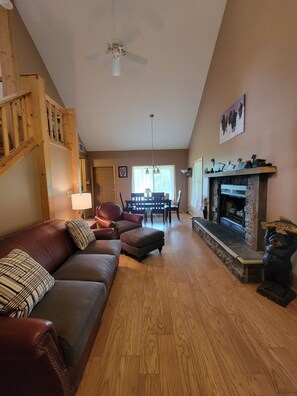 A living room with a fireplace, a wooden staircase, a dining area, and a sofa with cushions.