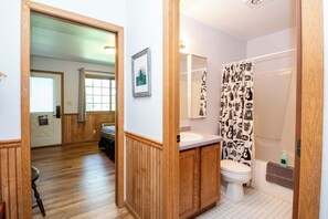 A bathroom with a wooden vanity, a toilet, a shower with a patterned curtain, and a framed picture on the wall.