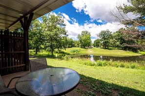 A covered outdoor seating area with a table and chairs, overlooking a pond and lush greenery.