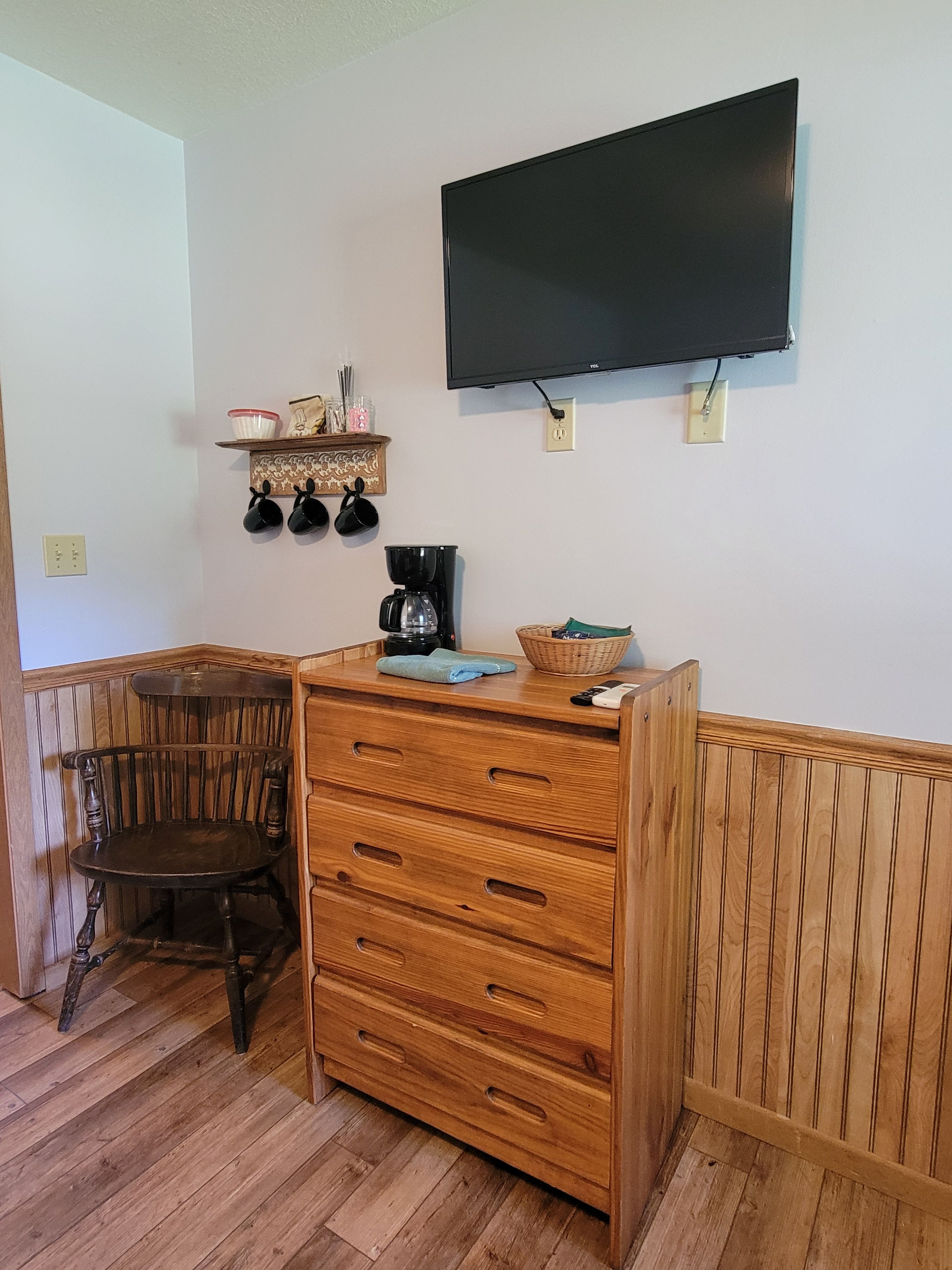 Une pièce comprenant une commode en bois, une chaise, une machine à café et un téléviseur fixé au mur.