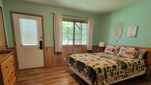 A bedroom with a wooden floor, a bed with a patterned quilt, a window with curtains, a door, and a wall with a dreamcatcher and a picture.
