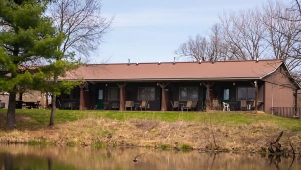 A building with a flat roof, a covered porch with chairs, and a pond in front.