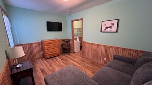 A room with a wooden floor, a television mounted on the wall, a framed picture, a dresser, and a chair.