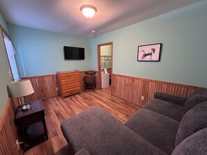 Ein Zimmer mit Holzboden, einem an der Wand befestigten Fernseher, einem gerahmten Bild, einem Schminktisch und einem Stuhl.