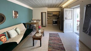 A living room with a sofa, coffee table, and a rug. There is a kitchen area with cabinets and appliances visible through an open door.